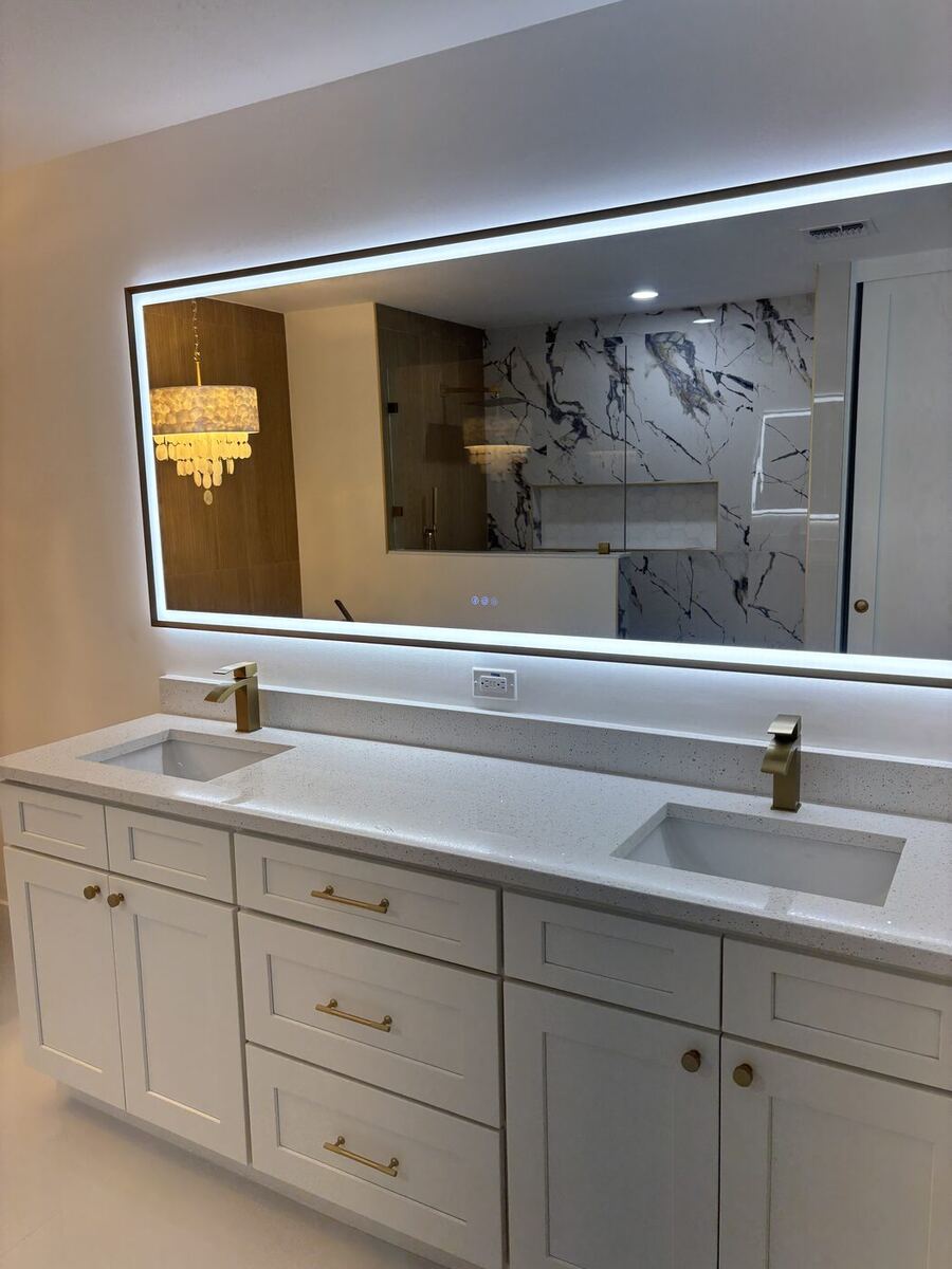 Double vanity with white shaker cabinets, gold faucets and a backlit mirror